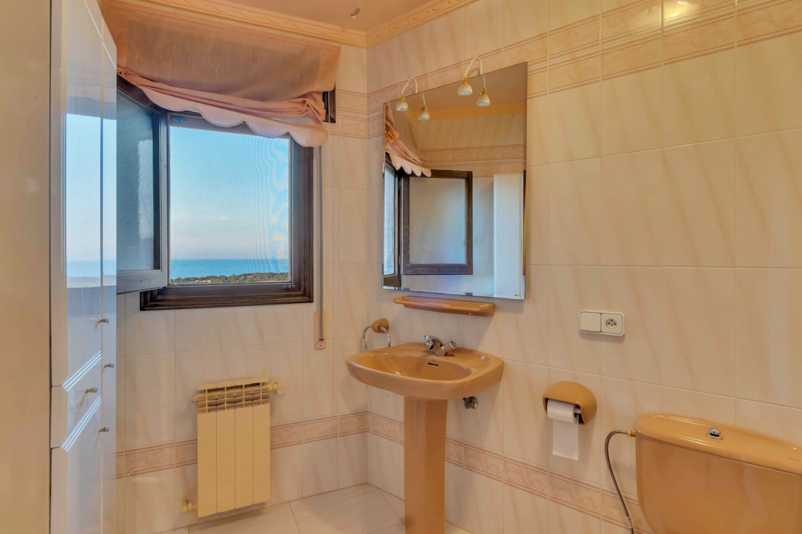Badezimmer mit beigefarbenen Keramikarmaturen, Säulenwaschbecken und Wandspiegel. Das Fenster bietet einen direkten Blick auf das Mittelmeer.