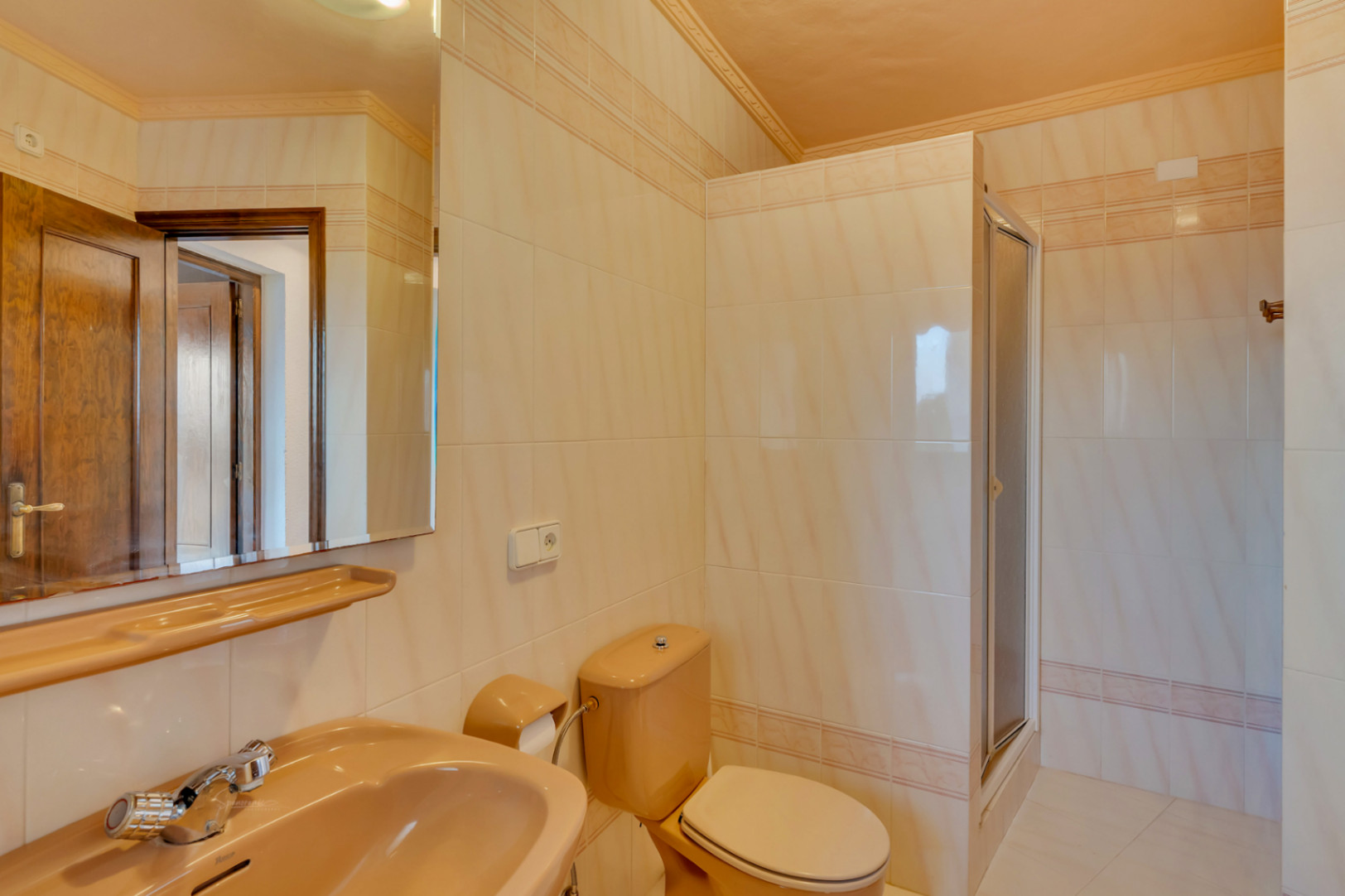 Badezimmer mit beigen Fliesen und Zierborte, ausgestattet mit pfirsichfarbenen Sanitäranlagen, großem Spiegel und einer begehbaren Dusche.