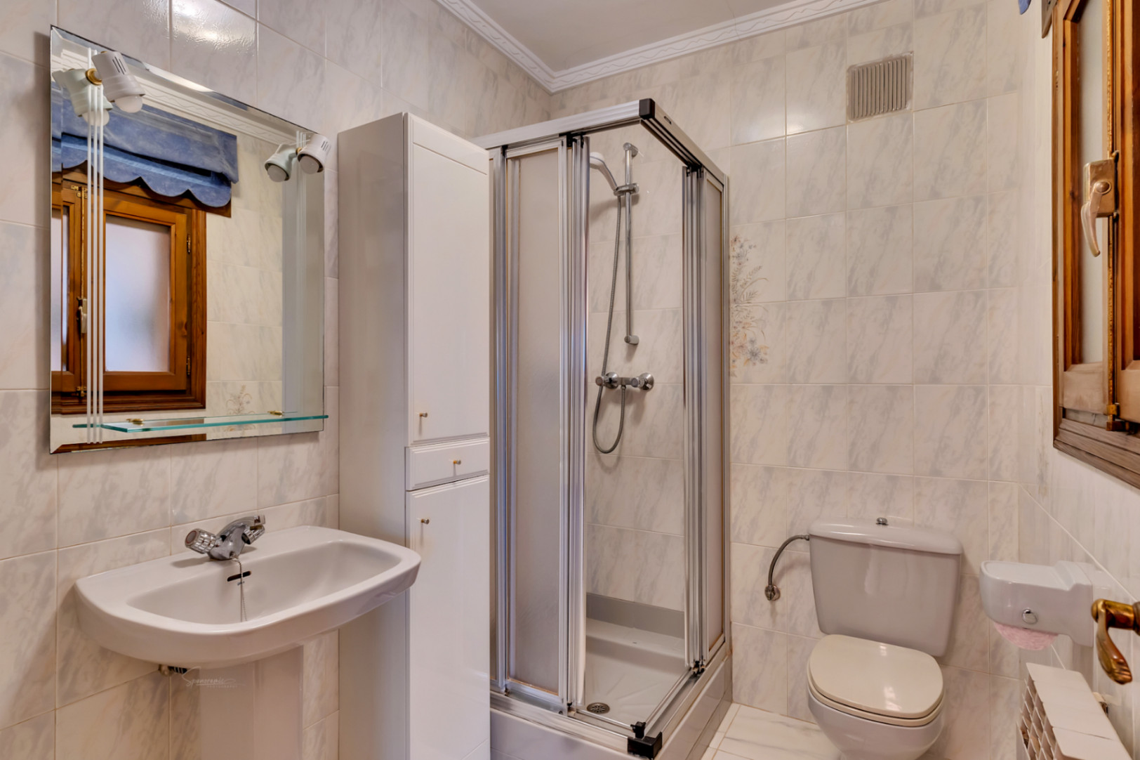 Badezimmer mit Standwaschbecken, hohem weißem Schrank und einer Eckduschkabine mit Schiebetüren vor hell gefliesten Wänden.
