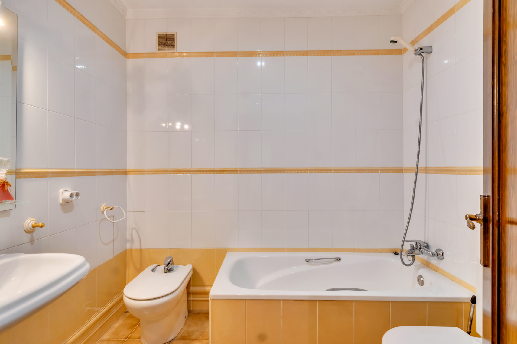 Funktionales Badezimmer mit Einbaubadewanne, Bidet und weißen Wandfliesen, die mit gelben Zierleisten und verchromten Armaturen versehen sind.