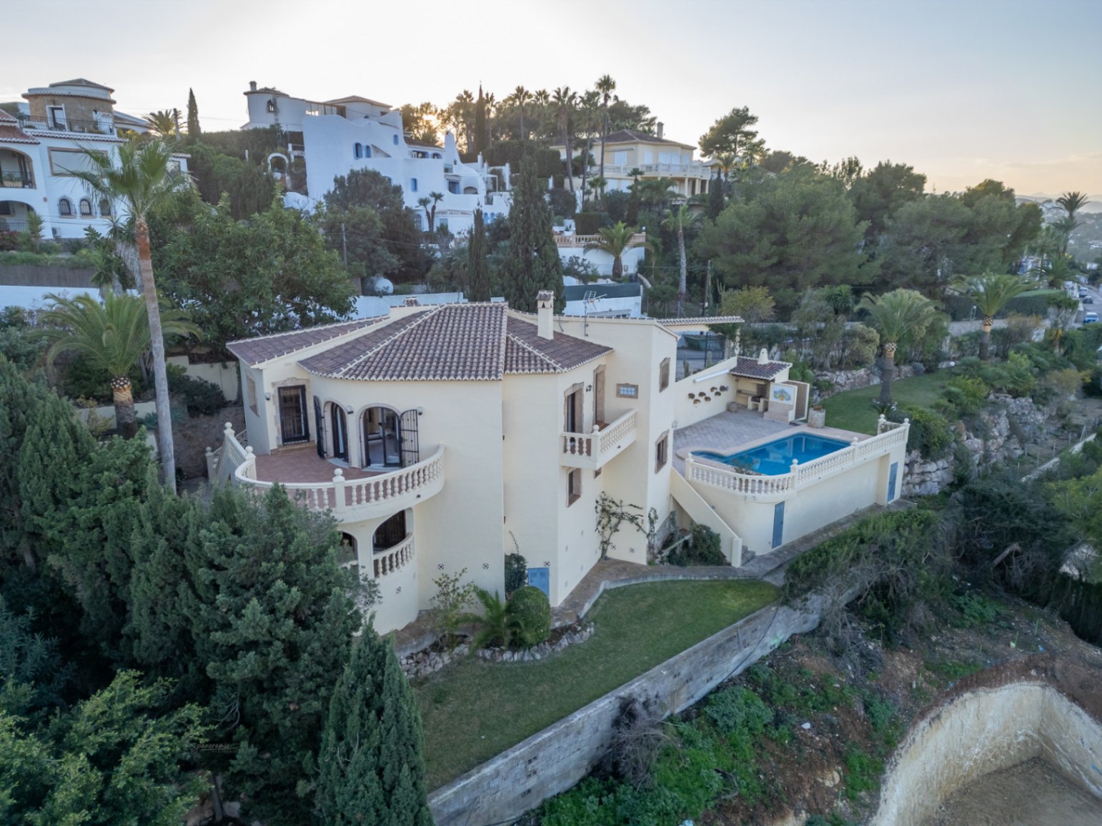 Luftaufnahme der mediterranen Villa mit Balustradenterrassen, rechteckigem Pool und Außengrillbereich inmitten gepflegter Gärten und Zypressen.