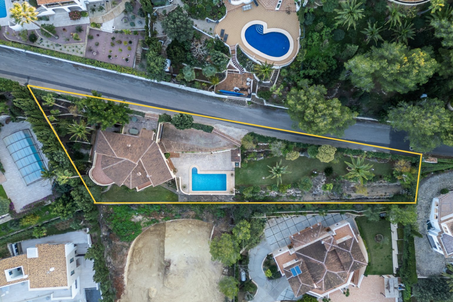Luftaufnahme des abgegrenzten Grundstücks mit L-förmigem Pool, mediterranem Garten inklusive Palmen und der Villa mit ihren charakteristischen Terrakotta-Ziegeldächern.