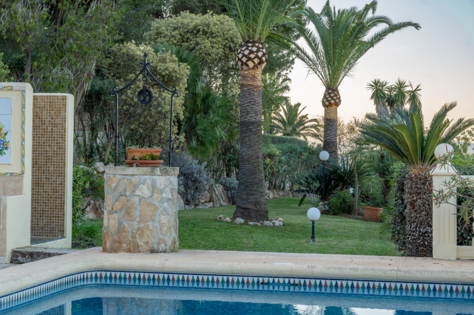 Mediterraner Garten mit Rasen, Palmen und einem dekorativen Steinbrunnen. Der Poolbereich bietet eine Außendusche mit Mosaikfliesen und Kugelleuchten.