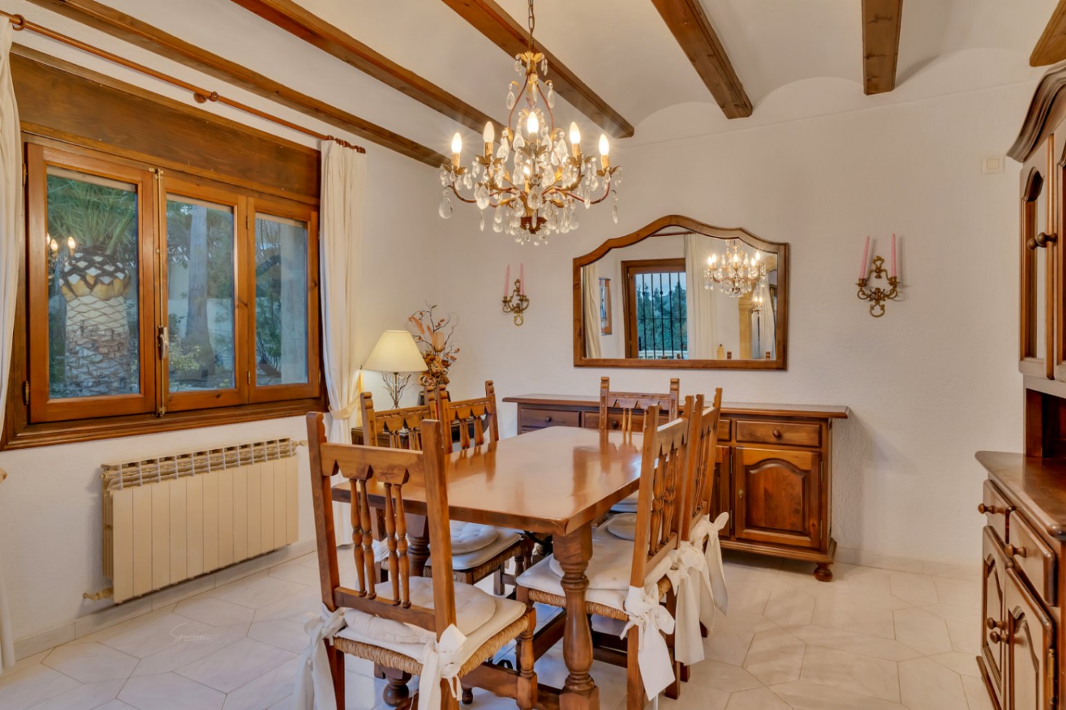 Rustikales Esszimmer mit sichtbaren Holzbalken, Massivholzmöbeln, einem Kristallleuchter und hellen sechseckigen Bodenfliesen.