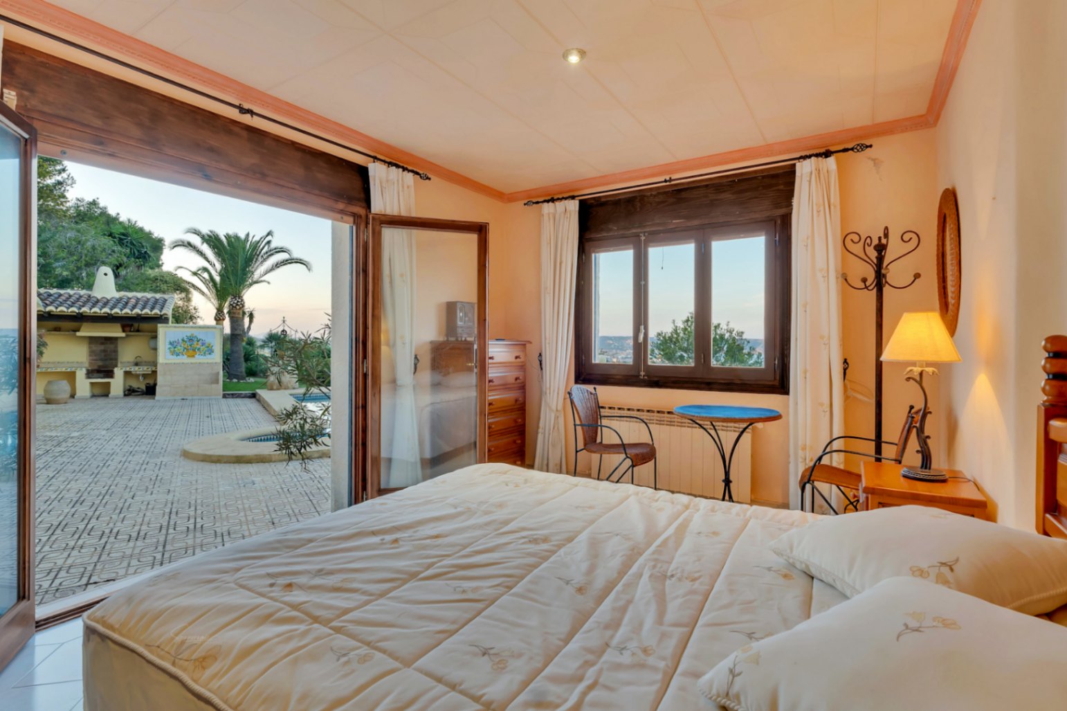 Schlafzimmer mit direktem Zugang zur Poolterrasse, dunklen Holzfensterrahmen und Blick auf den mediterranen Garten mit Grillplatz und Palmen.