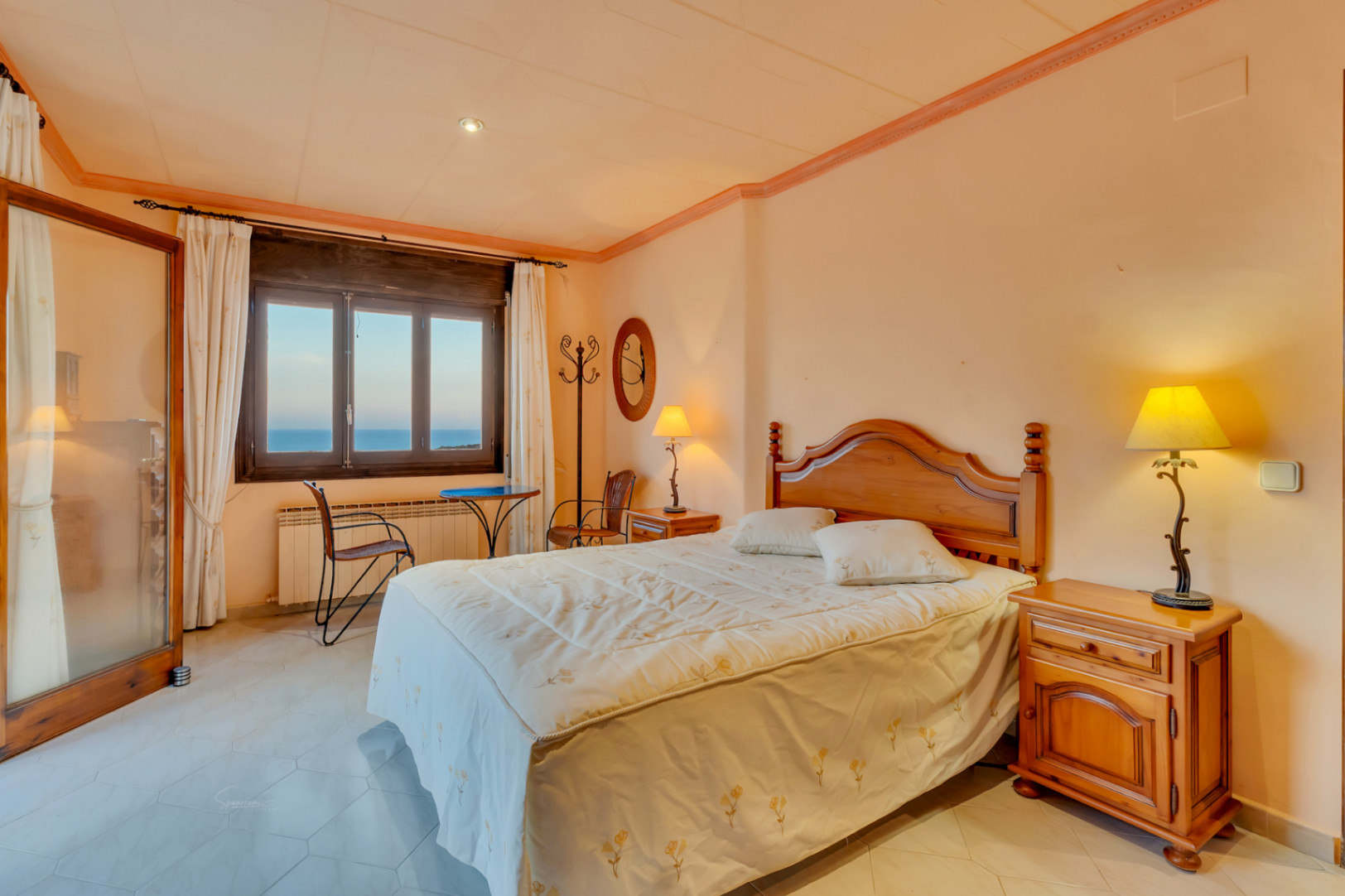 Schlafzimmer mit klassischen Holzmöbeln, Fliesenboden und Fenster mit Meerblick. Der Raum bietet eine warme Beleuchtung und eine kleine Sitzecke am Fenster.