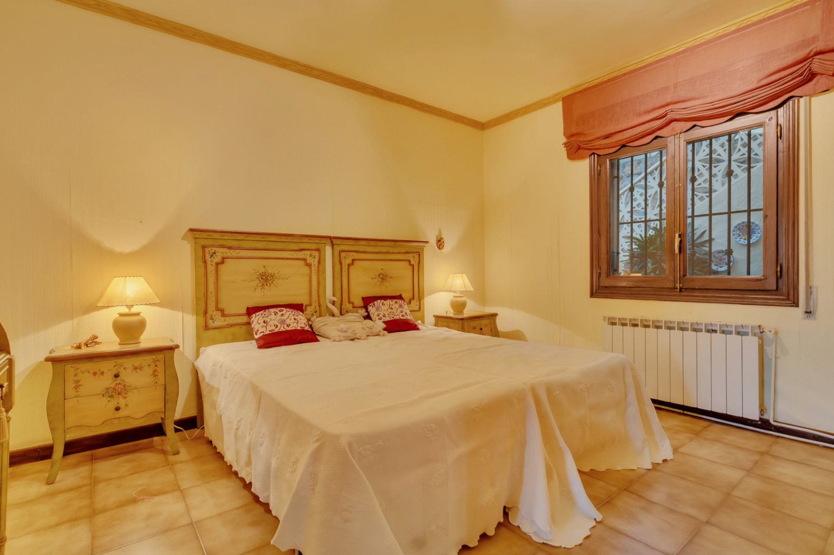 Schlafzimmer mit zwei Einzelbetten, verzierten Holzkopfteilen, Terrakotta-Fliesenboden, Heizkörper und Holzfenster mit Gittern.