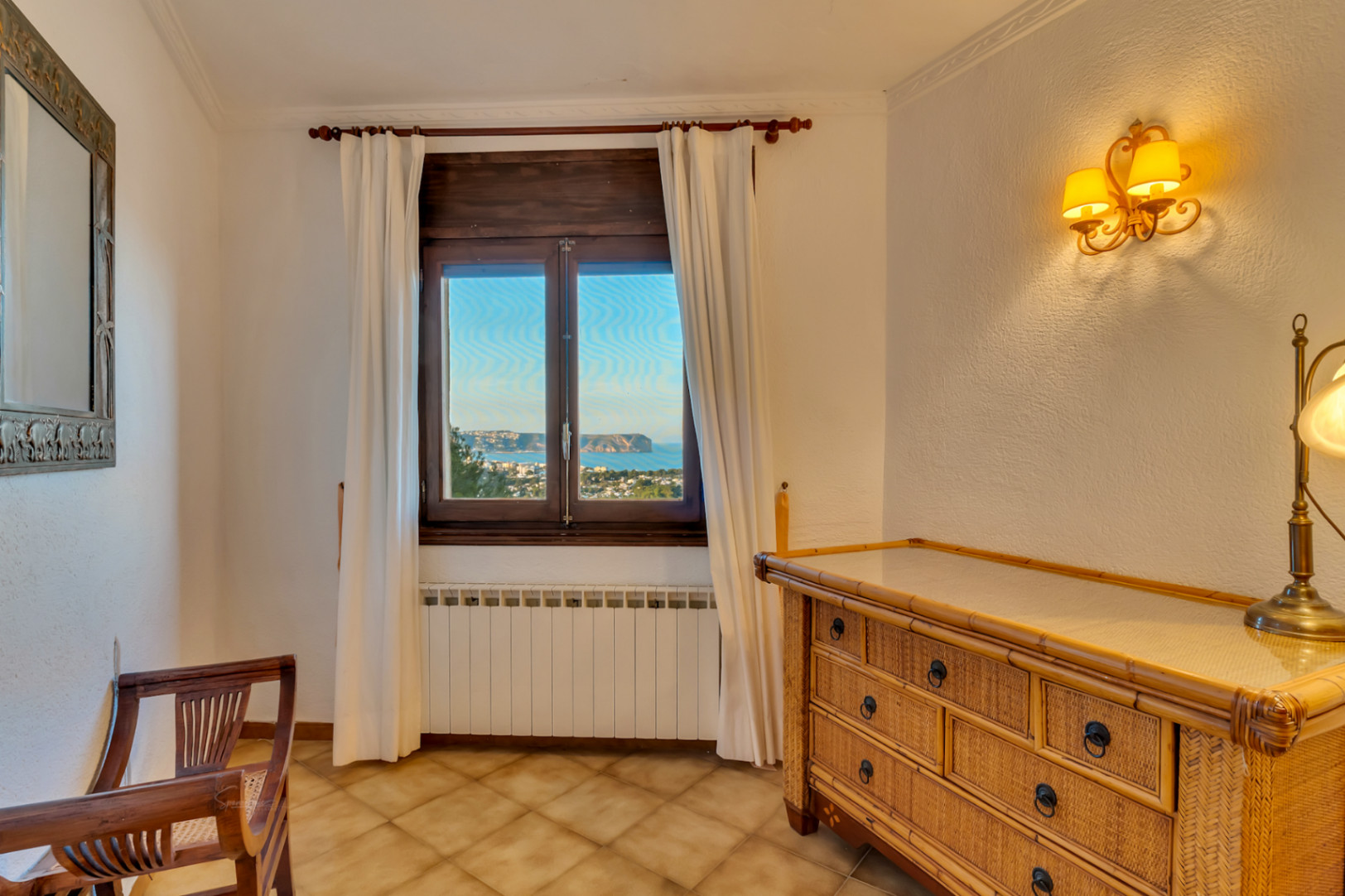 Schlafzimmerecke mit Flechtkommode, Holzstuhl und Fenster mit Blick auf das Meer und Cabo San Antonio. Ausgestattet mit Heizkörper und Wandbeleuchtung.