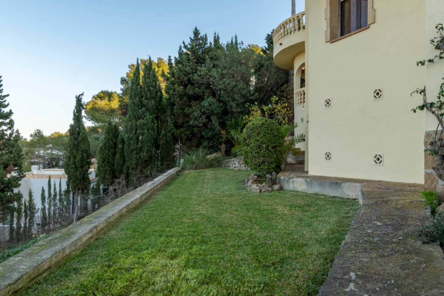 Seitlicher Garten mit gepflegtem Rasen, Steinmauern und Zypressen direkt an der hellen Fassade der Villa mit Balustradenbalkon.