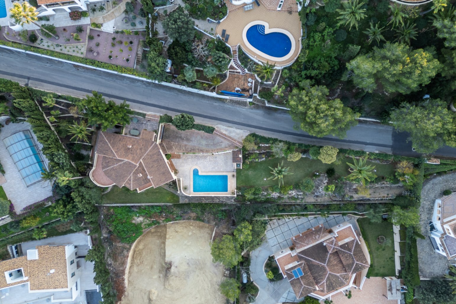 Senkrechte Luftaufnahme des Anwesens, die die Grundstücksaufteilung, den L-förmigen Pool, die gepflasterten Terrassen und den mediterranen Garten mit Palmen zeigt.