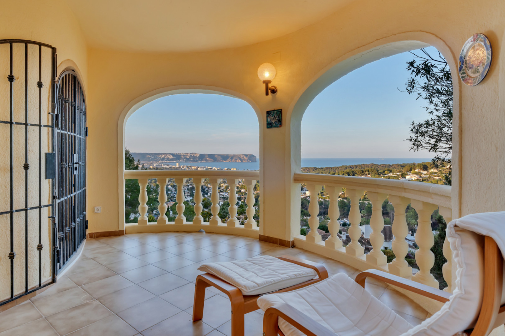 Überdachte Bogenterrasse mit klassischer Balustrade und Panoramablick auf die Bucht von Jávea. Ausgestattet mit Liegestuhl, Fliesenboden und dekorativem Schmiedeeisentor.