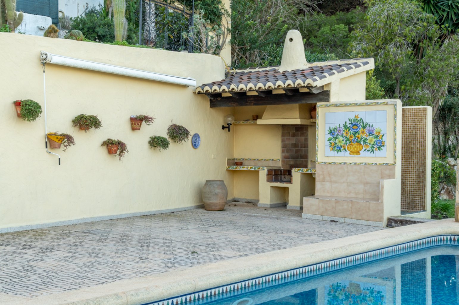 Überdachter Grillbereich am Pool mit dekorativem Fliesenmosaik, gemustertem Steinboden und einer Außenbrause im mediterranen Stil.
