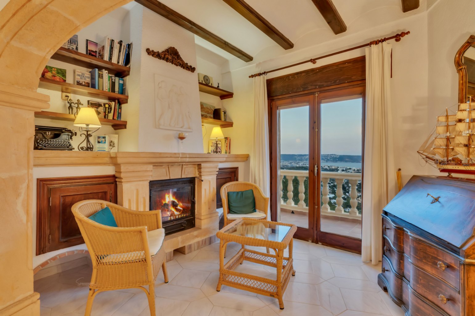 Wohnzimmer mit Tosca-Steinkamin, Holzbalken und klassischem Sekretär. Die Glastüren führen zum Balkon mit Blick auf die Bucht von Jávea.