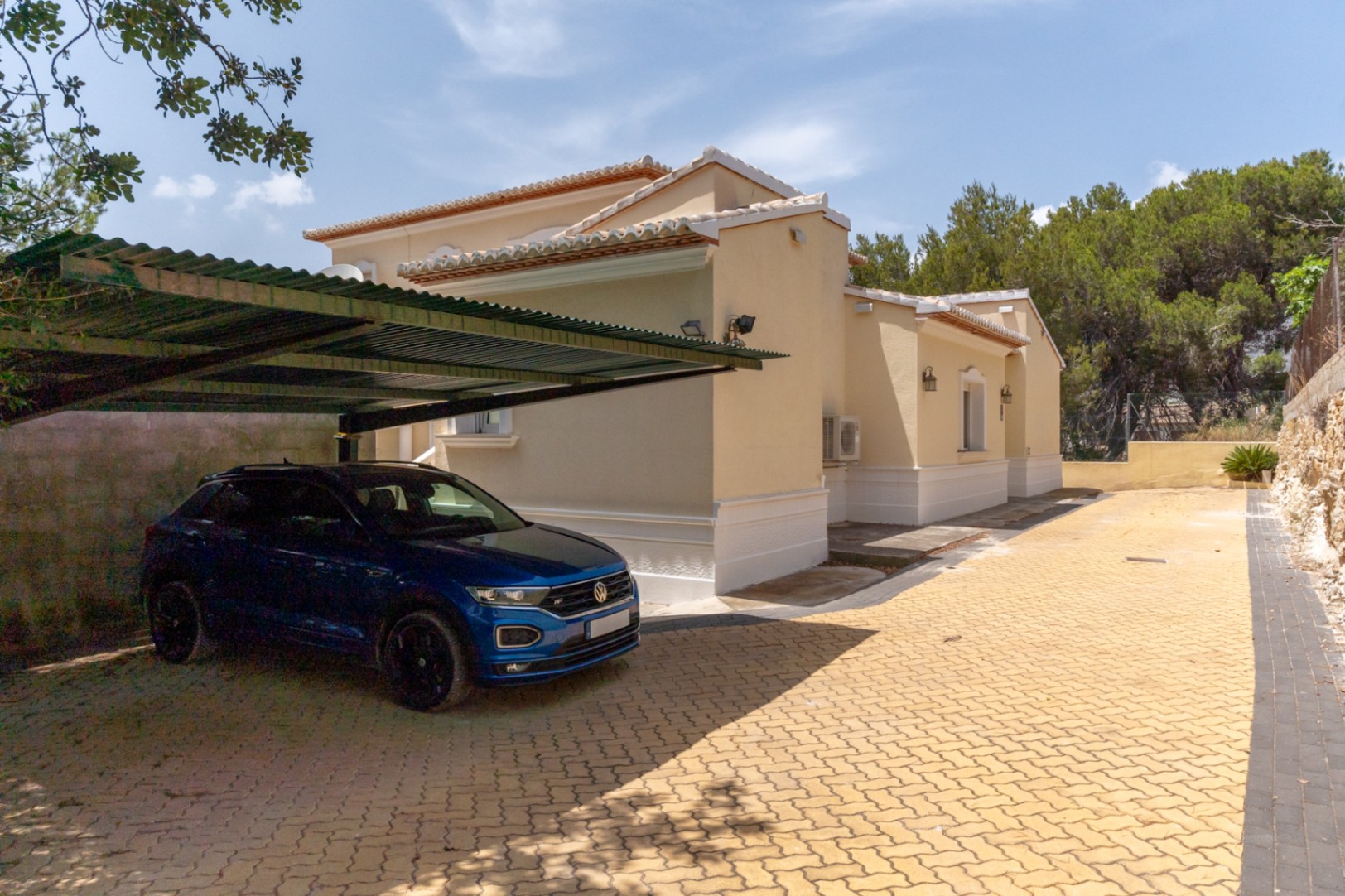 Außenparkplatz mit Metall-Carport und gepflasterter Einfahrt neben der beigefarbenen Fassade der Villa mit dekorativen weißen Sockeln.