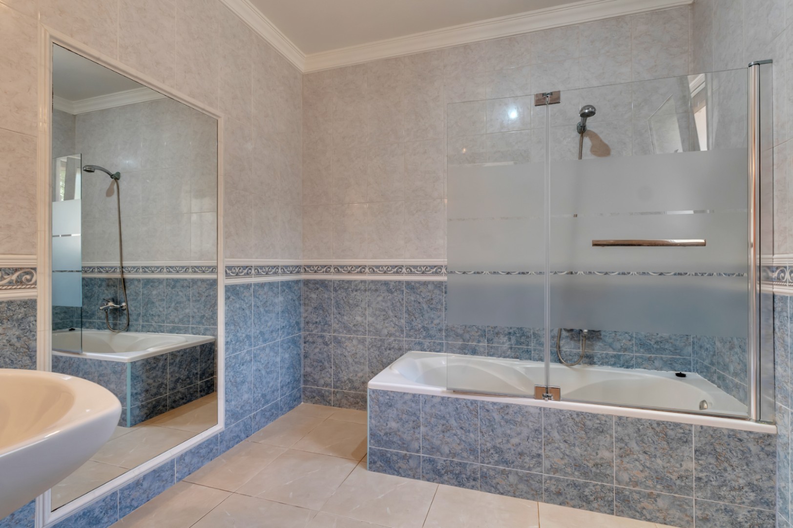 Badezimmer mit Einbaubadewanne, teilmattierter Glastrennwand und blau-beige gefliesten Wänden mit dekorativer Bordüre.
