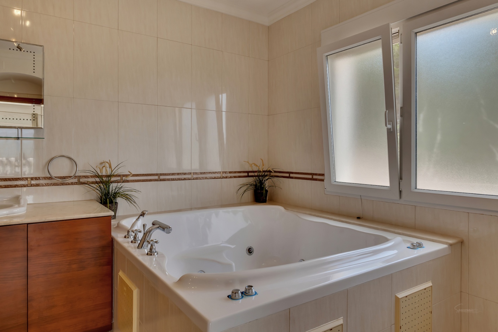 Badezimmer mit eingebauter Hydromassage-Badewanne, cremefarbenen Wandfliesen mit dekorativer Bordüre und einem doppelt verglasten Milchglasfenster für Privatsphäre.