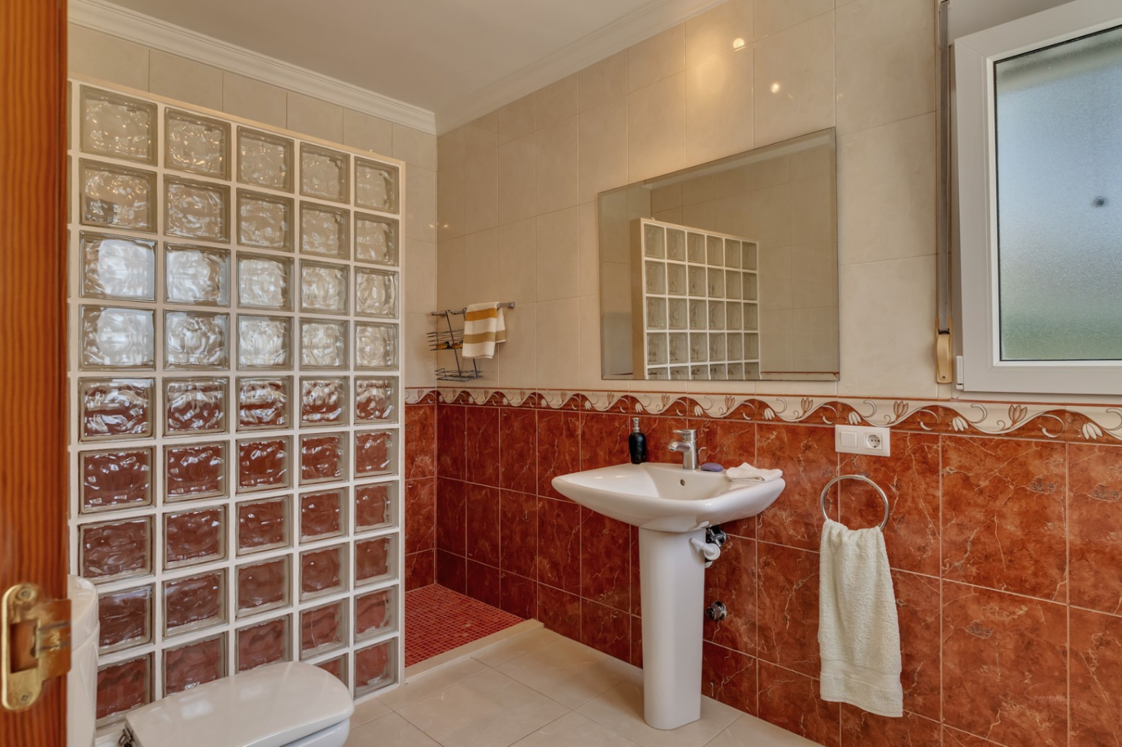 Badezimmer mit Standwaschbecken, großem Spiegel und einer Dusche mit Glasbausteinwand. Die Wände sind mit rötlichen Marmor-Effekt-Fliesen und einer Zierbordüre im klassischen Stil gestaltet.
