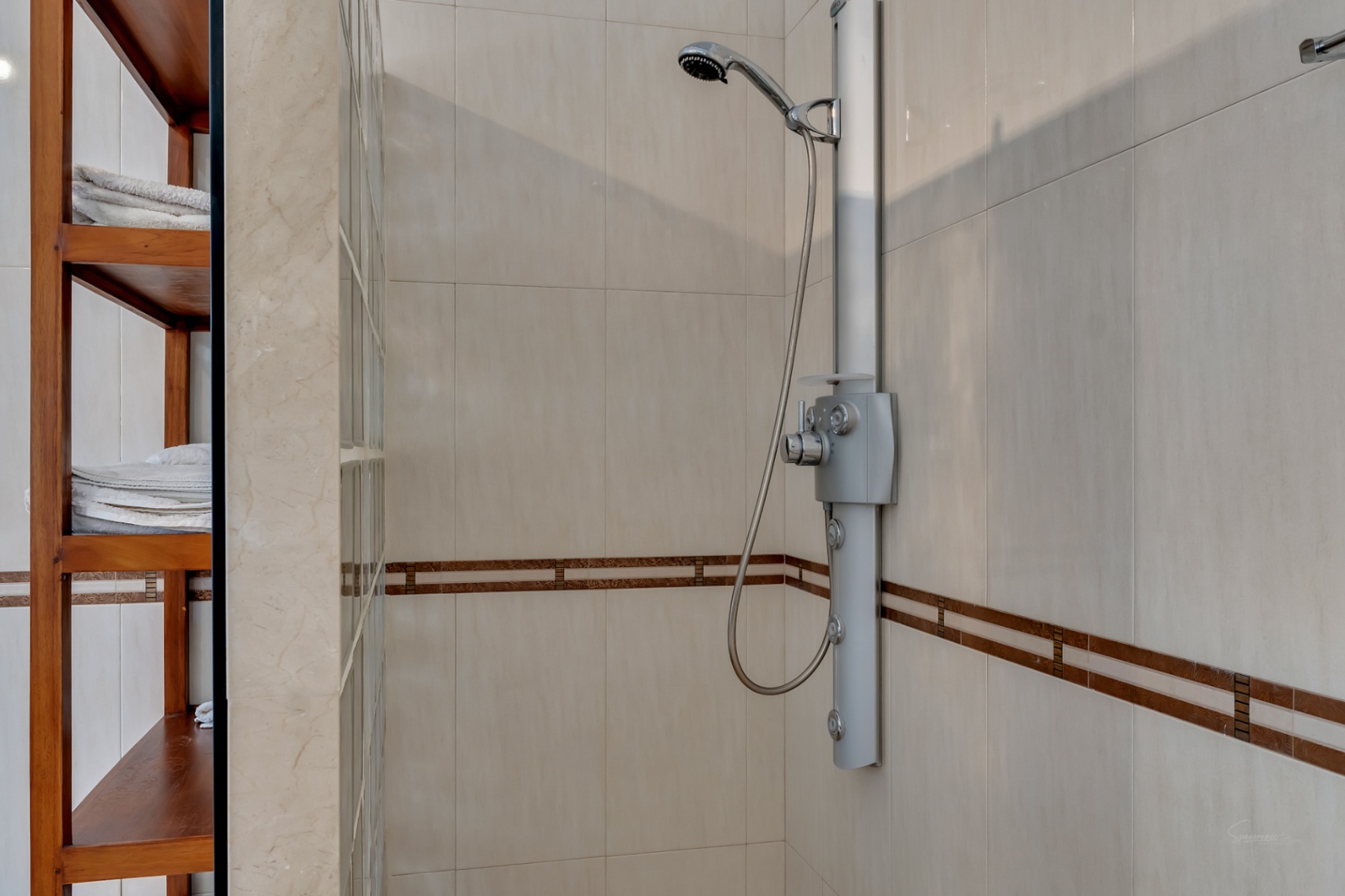 Duschbereich mit Hydromassage-Säule, cremefarbenen Fliesen mit dekorativer Bordüre und einem Holzregal neben einer Wand aus Glasbausteinen.