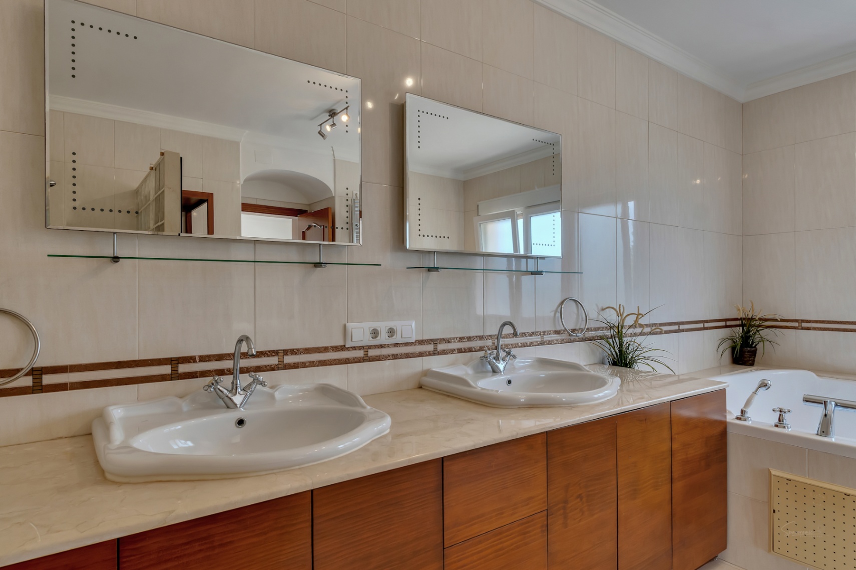 En-Suite-Badezimmer mit Doppelwaschbecken auf Marmorplatte, Holzschränken und hintergrundbeleuchteten Spiegeln neben der Hydromassage-Badewanne.