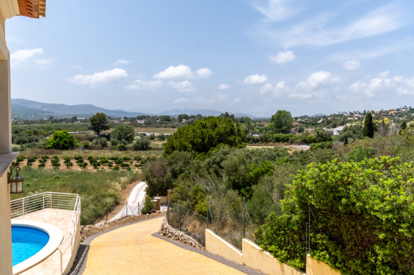 Erhöhter Blick auf die gepflasterte Auffahrt, den geschwungenen Pool mit weißem Geländer und die weite Aussicht über das Tal von Jávea mit seinen Orangenhainen.