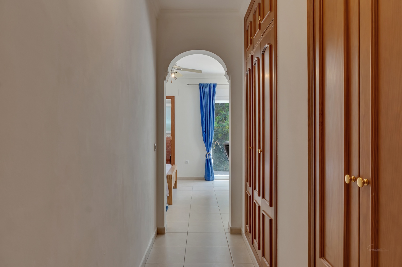 Flur mit großen Einbauschränken aus Holz und Keramikboden, der durch einen Rundbogen zu einem hellen Schlafzimmer mit Deckenventilator und blauen Vorhängen führt.