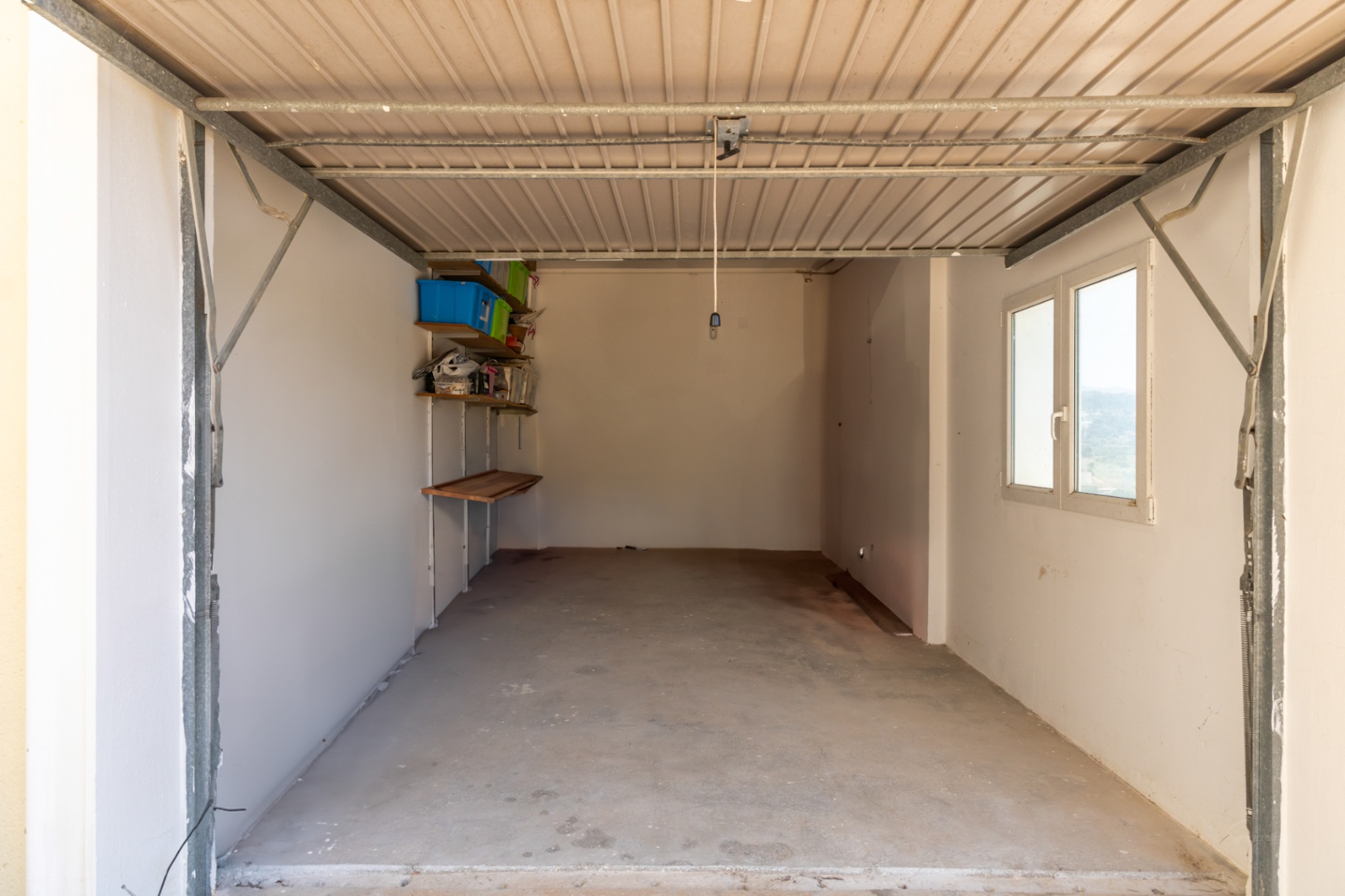 Geschlossene Garage mit Betonboden, weißen Wänden und Seitenfenster. Ausgestattet mit Wandregalen für zusätzlichen Stauraum.