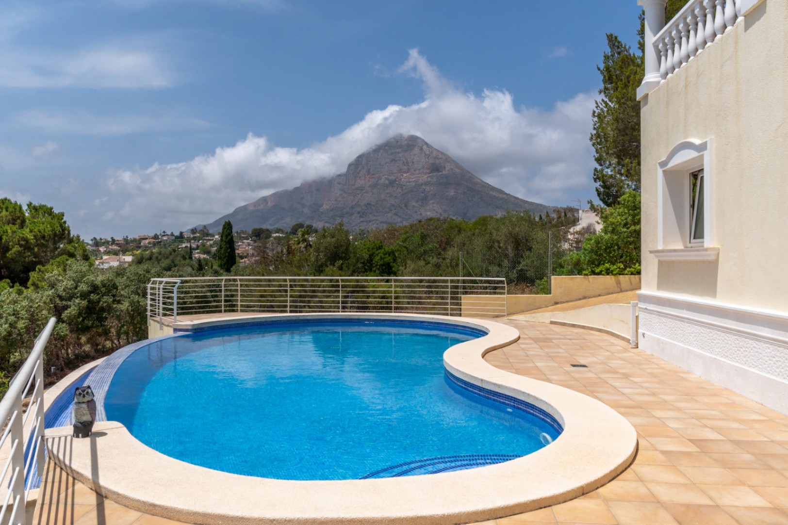 Geschwungener Infinity-Pool mit blauen Mosaikfliesen und Panoramablick auf den Berg Montgó, umgeben von einer gefliesten Terrasse mit Metallgeländer.