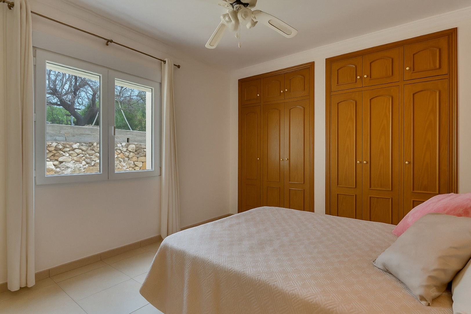 Helles Schlafzimmer mit Einbauschränken aus Holz, Deckenventilator und Fenster mit Blick auf den Garten und eine Natursteinmauer.