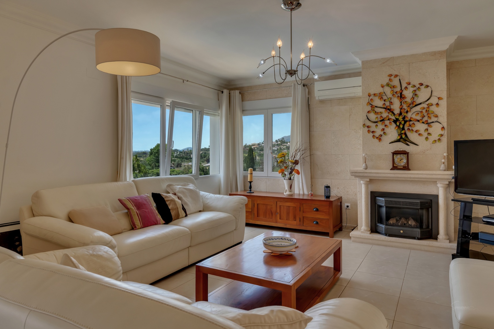 Helles Wohnzimmer mit weißen Ledersofas, einem Steinkamin mit Holzofen und Fenstern mit Panoramablick auf das Tal und die Landschaft.