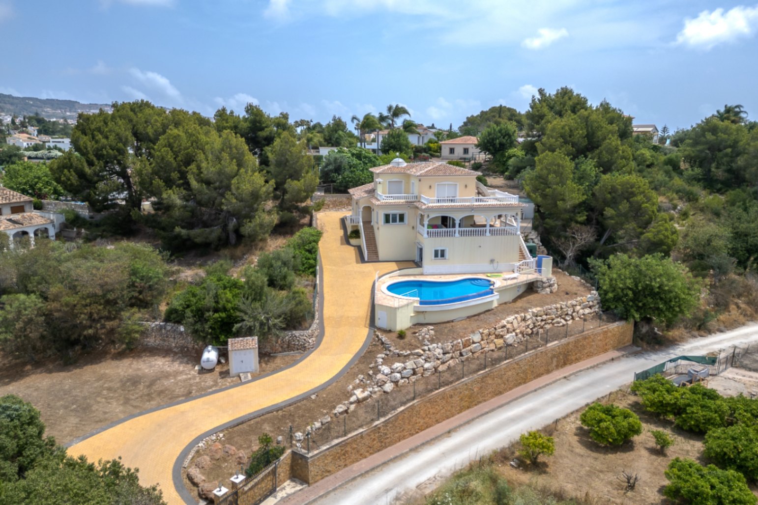 Luftaufnahme der erhöht gelegenen Villa mit geschwungenem Infinity-Pool, weitläufigen Terrassen und einer langen gepflasterten Auffahrt inmitten von Pinien.