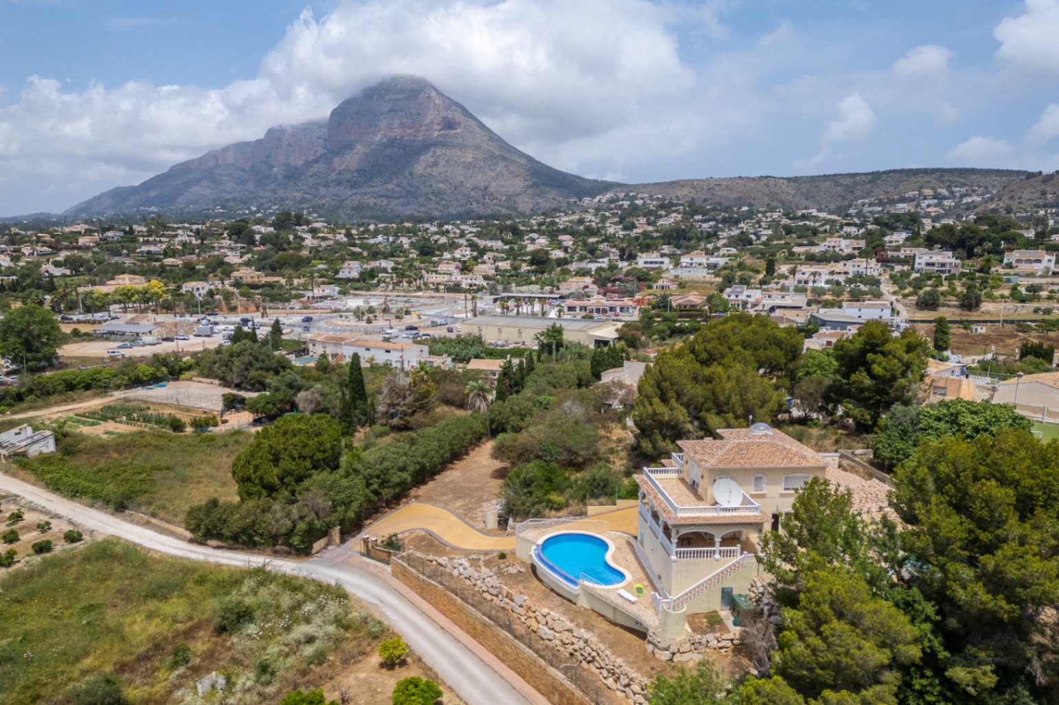 Luftaufnahme der Villa mit geschwungenem Infinity-Pool und erhöhter Lage, die einen weiten Blick über das Tal bis hin zum Berg Montgó bietet.