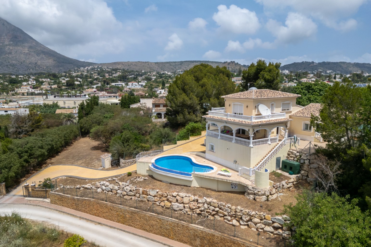 Luftaufnahme der Villa mit geschwungenem Pool, naya mit Rundbögen und Terrassen mit Balustraden, eingebettet in die Landschaft mit Blick auf den Montgó.