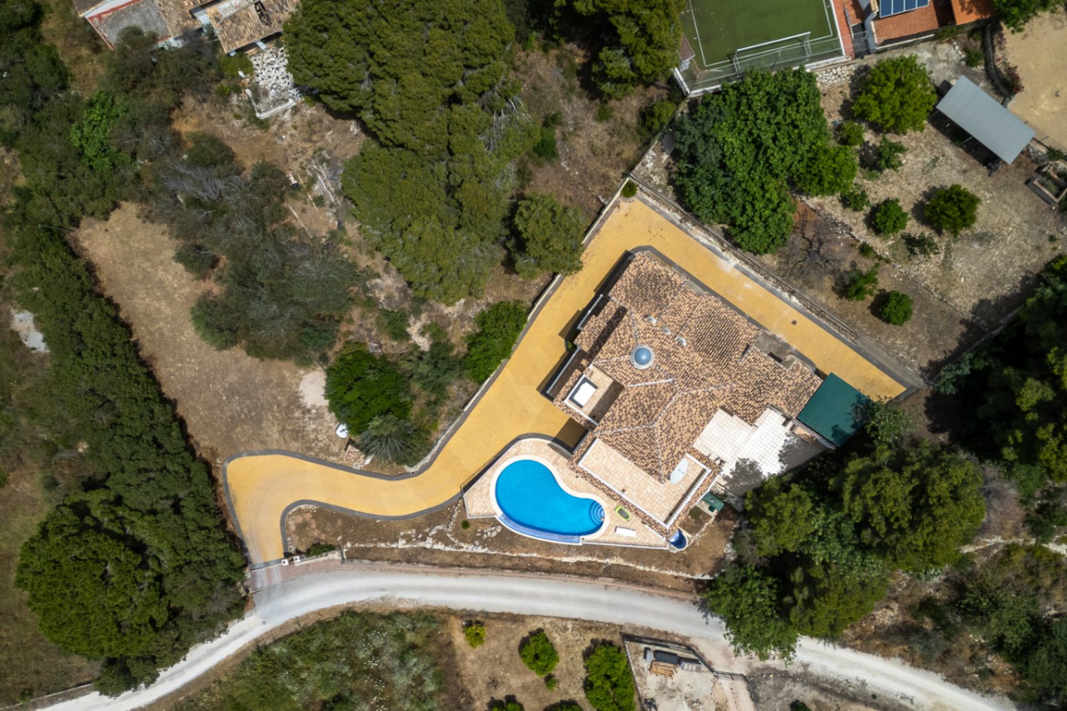 Luftaufnahme der Villa mit Ziegeldach und zentraler Glaskuppel, einem geschwungenen Infinity-Pool und einer gepflasterten Auffahrt inmitten mediterraner Natur.