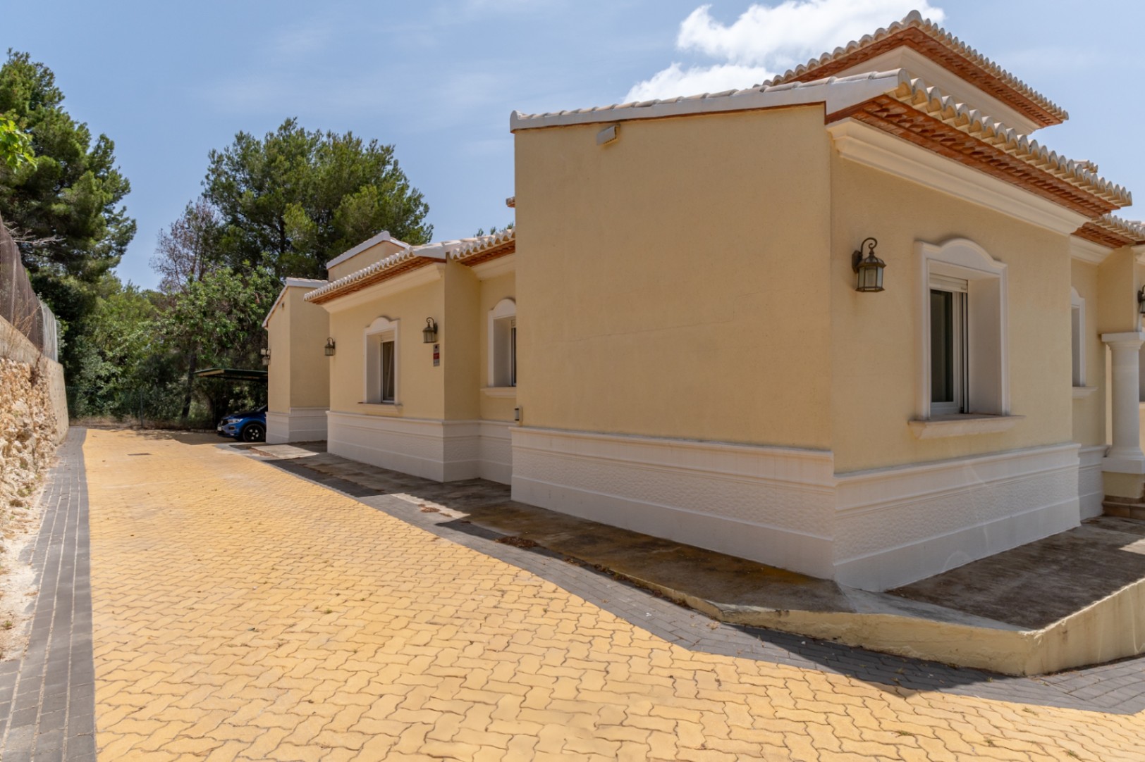 Ockerfarbene Außenfassade mit weißen Zierleisten, gepflasterte Einfahrt und überdachter Parkplatz, umgeben von mediterranen Pinien.