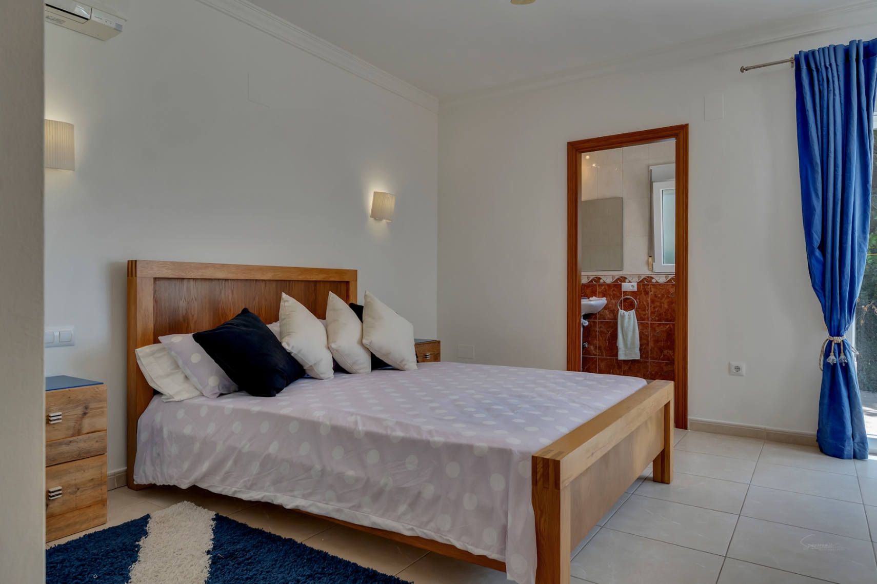 Schlafzimmer mit Holzbett, Fliesenboden und En-Suite-Badezimmer mit rötlichen Wandfliesen. Ausgestattet mit Klimaanlage und dezenten Wandleuchten.