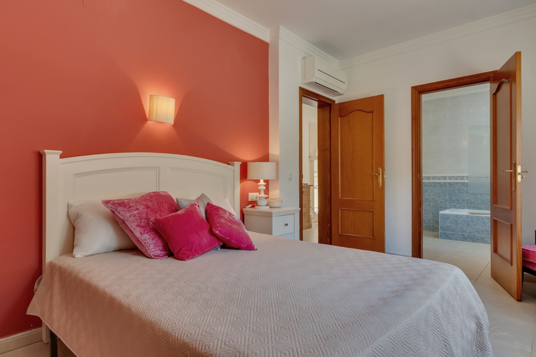 Schlafzimmer mit roter Akzentwand, weißem Kopfteil und Klimaanlage. Eine Massivholztür führt zum En-Suite-Badezimmer mit blauen Fliesen und eingebauter Badewanne.