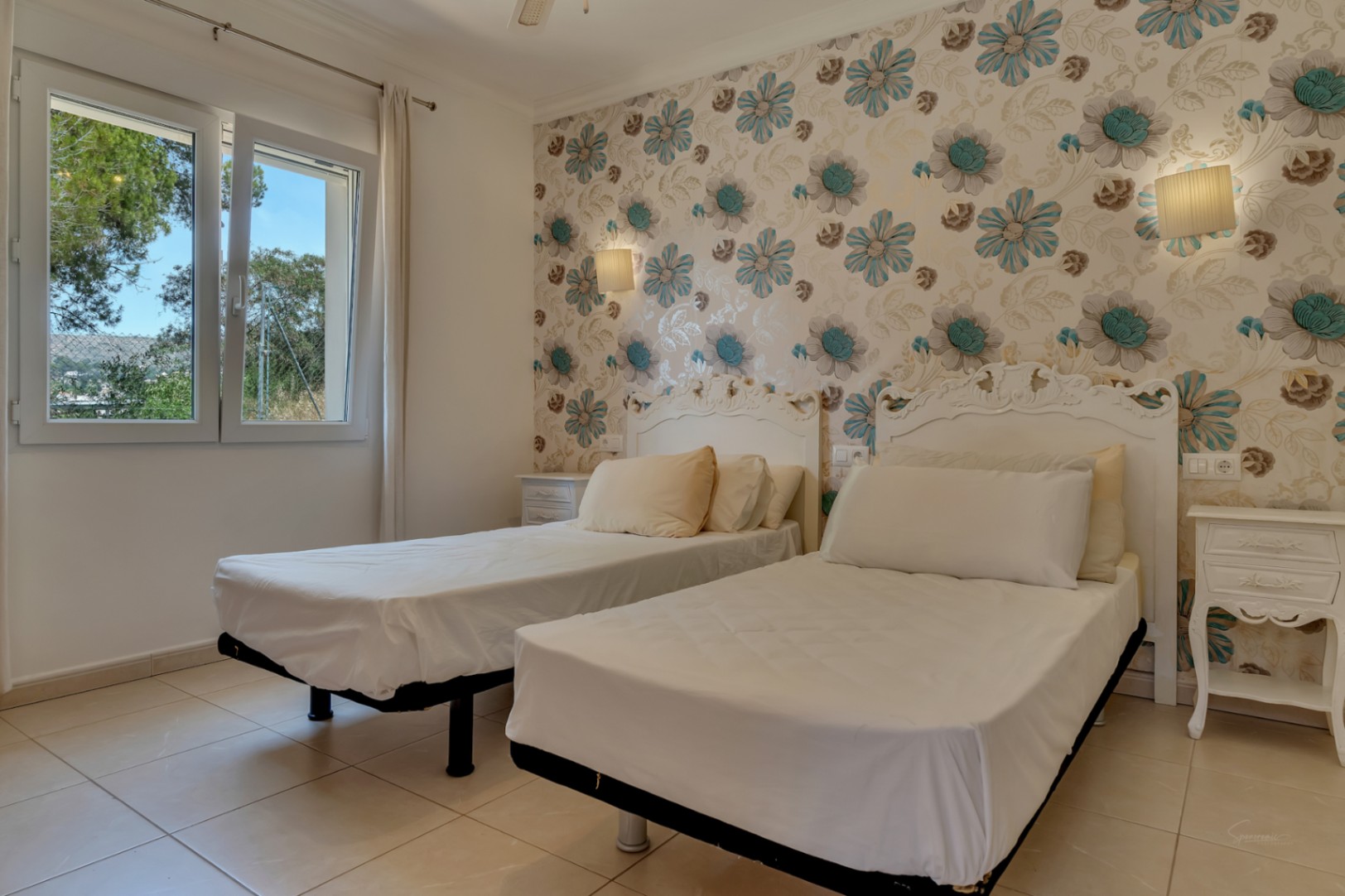 Schlafzimmer mit zwei Einzelbetten, verzierten weißen Kopfteilen und floraler Mustertapete. Mit Fliesenboden und einem Fenster mit Blick auf die umliegende Landschaft.
