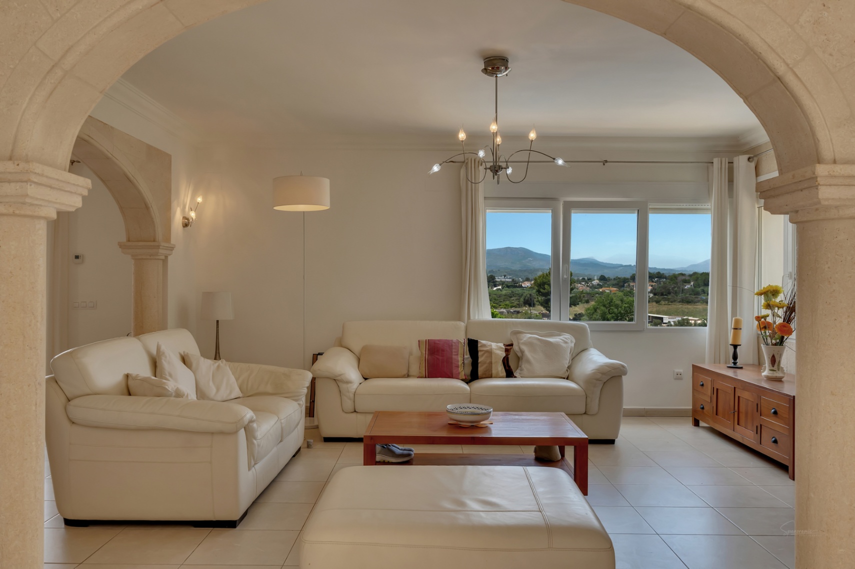 Wohnzimmer mit weißen Ledersofas und traditionellen Steinbögen, das durch ein großes Fenster einen Panoramablick auf das Tal und die Berge bietet.