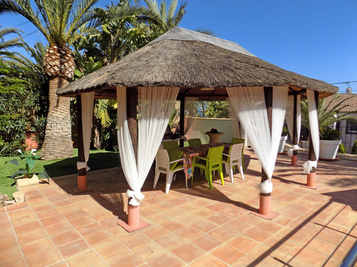 Außenpavillon mit Reetdach und weißen Vorhängen auf einer Terrakotta-Terrasse, eingebettet in einen Garten mit Palmen und Essbereich.