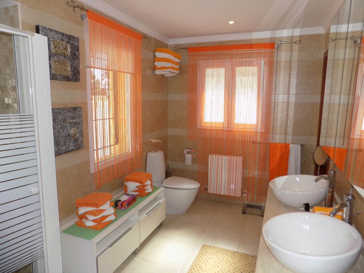 Komplettes Badezimmer mit Doppelwaschbecken, cremefarbenen Fliesenwänden und natürlichem Licht durch zwei Fenster. Ausgestattet mit Heizkörper und modernen Sanitärobjekten.