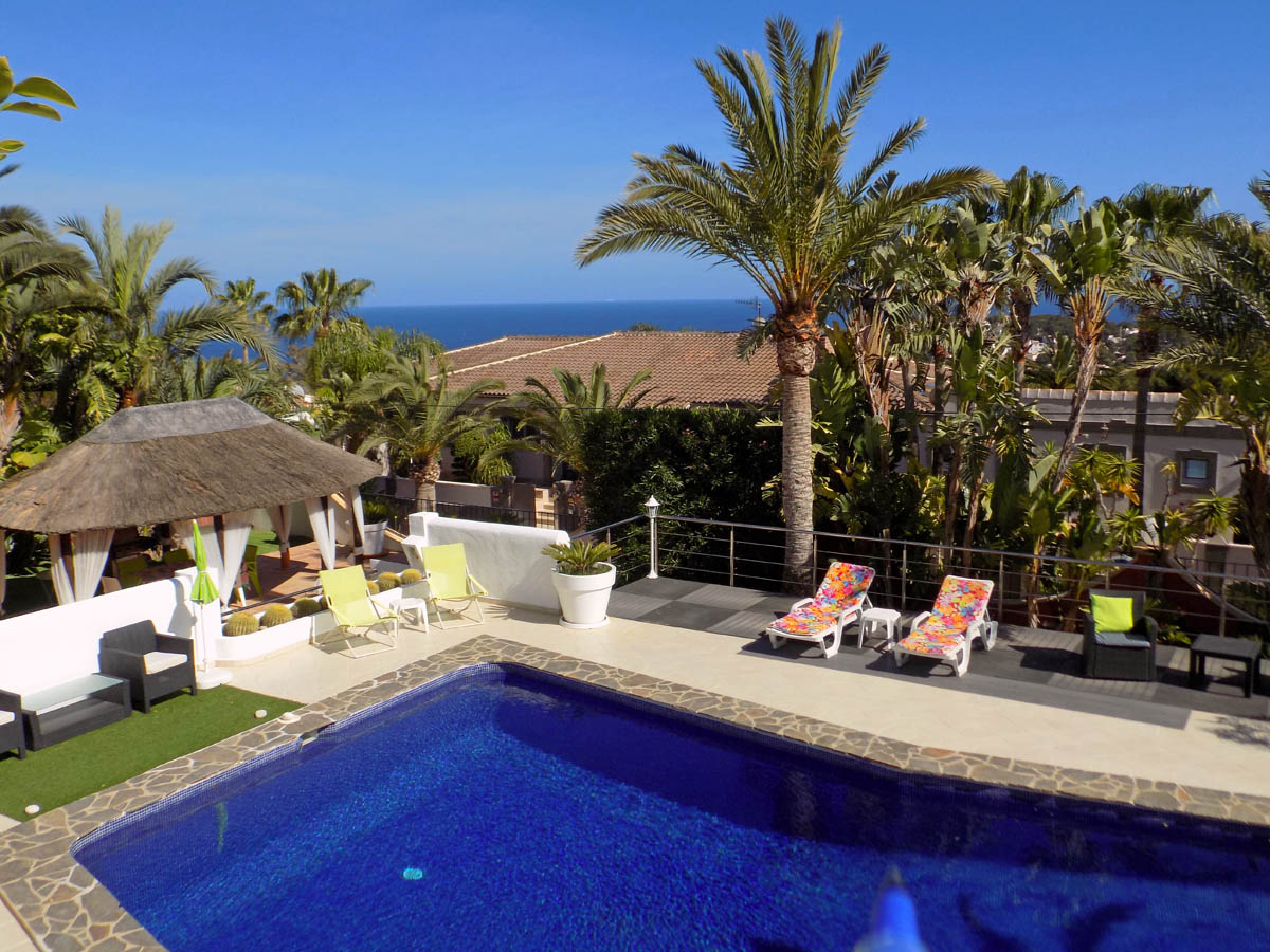 Poolbereich mit Natursteineinfassung, reetgedeckter Chill-out-Ecke und Panoramablick auf das Mittelmeer, umgeben von üppiger mediterraner Vegetation.