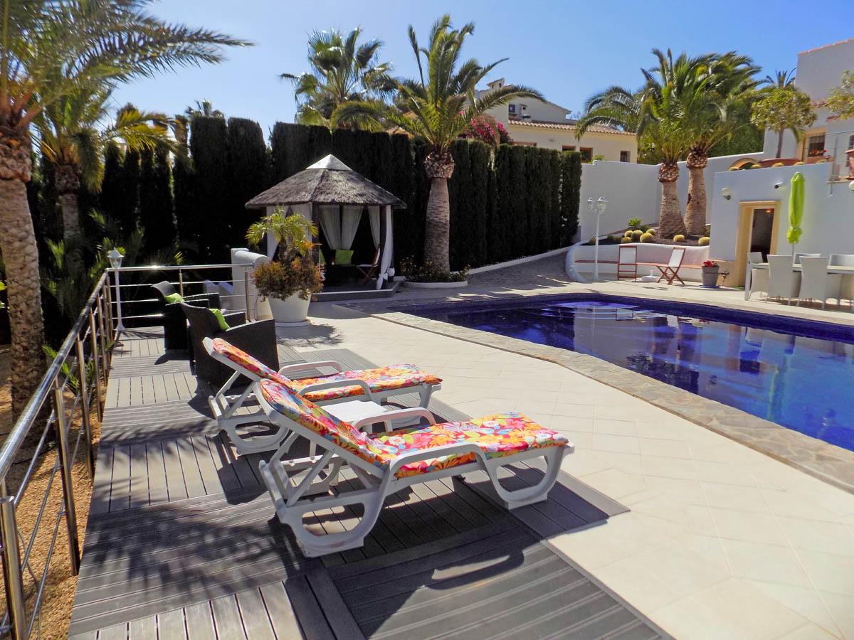 Poolbereich mit Stein- und Verbundstoff-Terrasse, Sonnenliegen, einem reetgedeckten Pavillon und mediterraner Bepflanzung mit hohen Palmen.