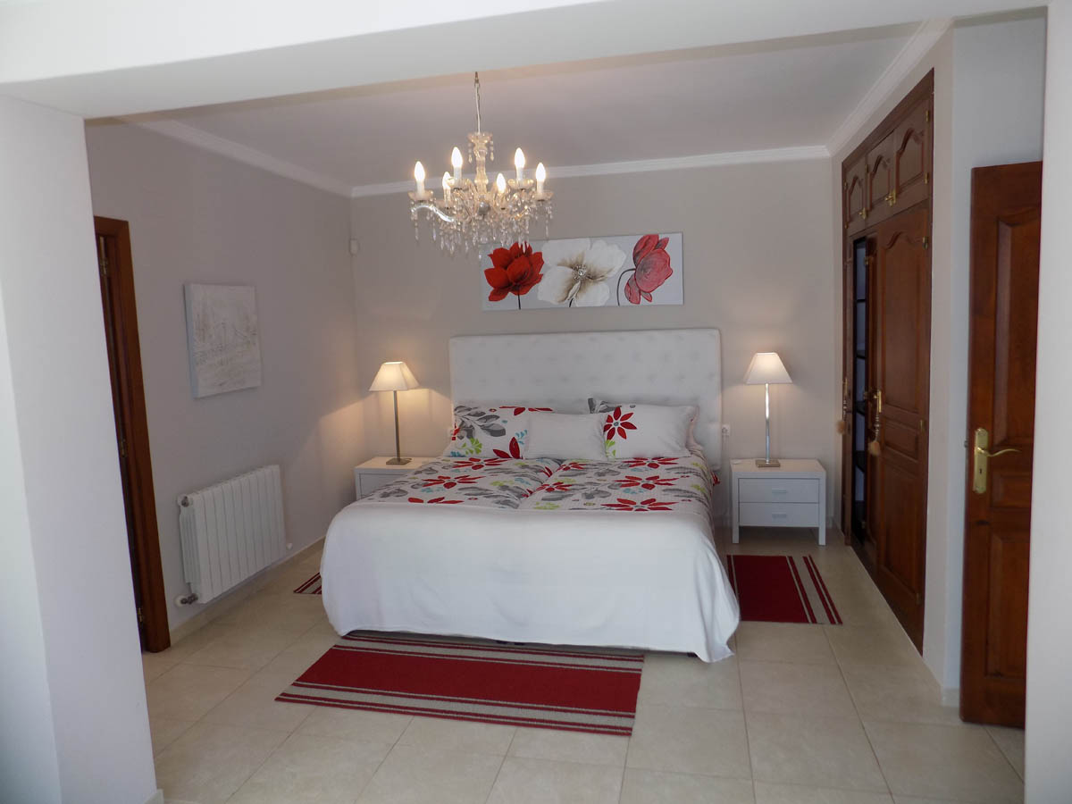 Schlafzimmer mit hölzernen Einbauschränken, weißem Polsterkopfteil, Kristallkronleuchter und hellen Fliesenböden mit Heizkörper.
