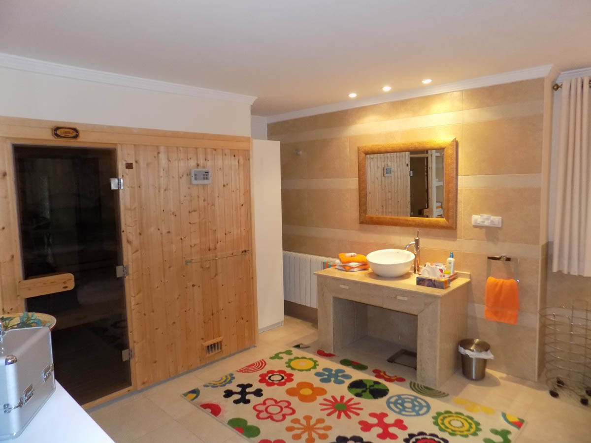 Wellnessbereich mit integrierter Holzsauna, Naturstein-Waschtisch mit Aufsatzbecken und warm getönten Fliesenwänden mit Deckenspots.
