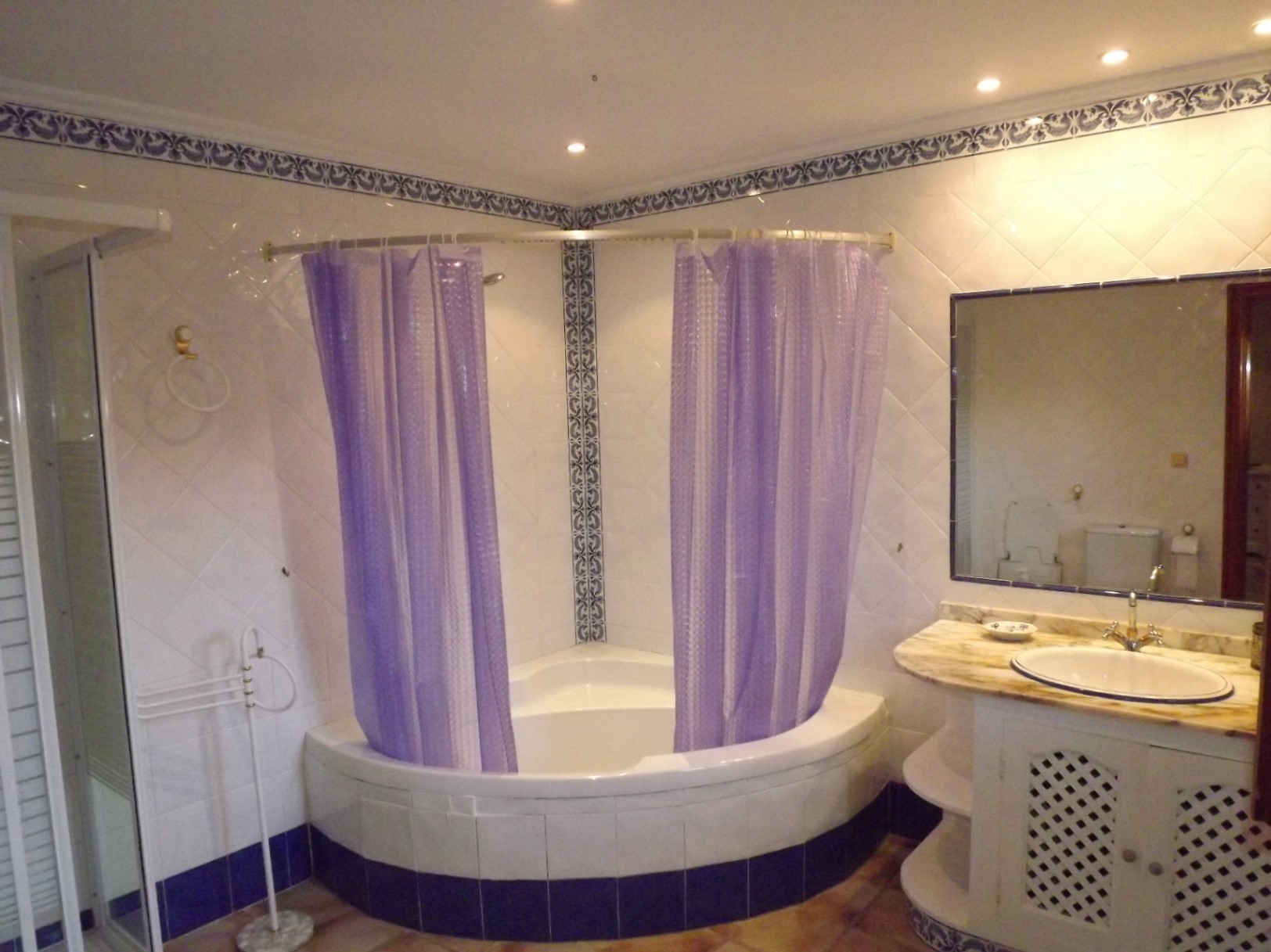 Badezimmer mit Eckbadewanne und violetten Vorhängen, Marmor-Waschtisch mit Gittertüren und weißen Fliesen mit blauen Zierbordüren.