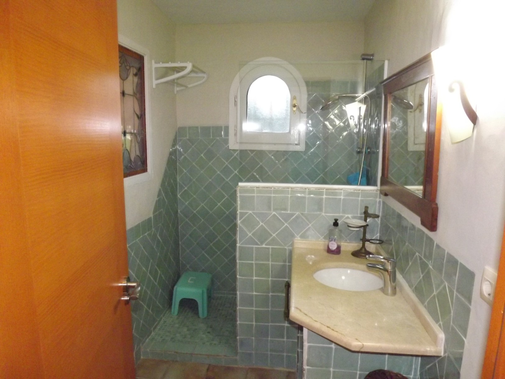 Badezimmer mit grünen Fliesen, Naturstein-Waschtisch und begehbarer Dusche mit Rundbogenfenster und Glastrennwand. Mit dekorativem Buntglasfenster.