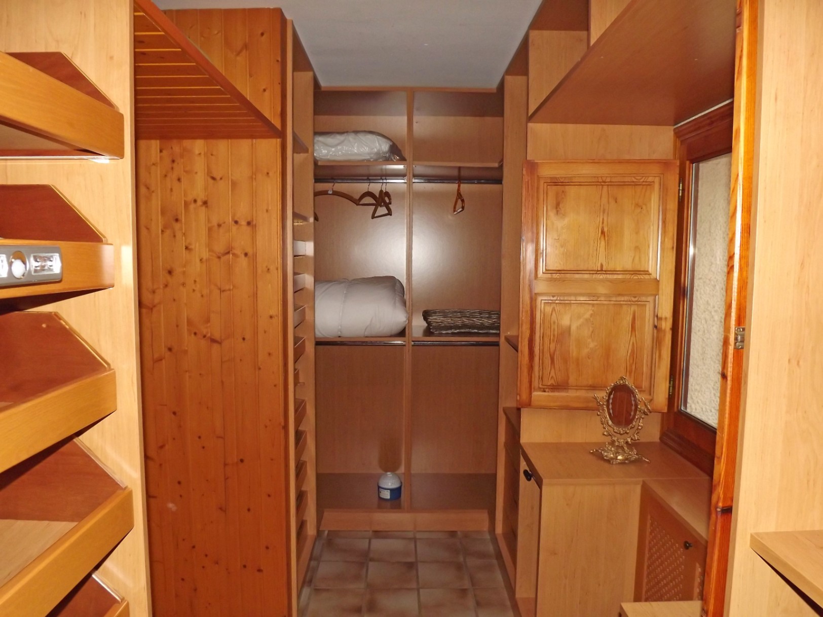 Begehbarer Kleiderschrank mit maßgefertigten Holzregalen, Kleiderstangen und Fliesenboden. Ein Fenster mit Holzschlagladen sorgt für natürliches Licht und Belüftung.