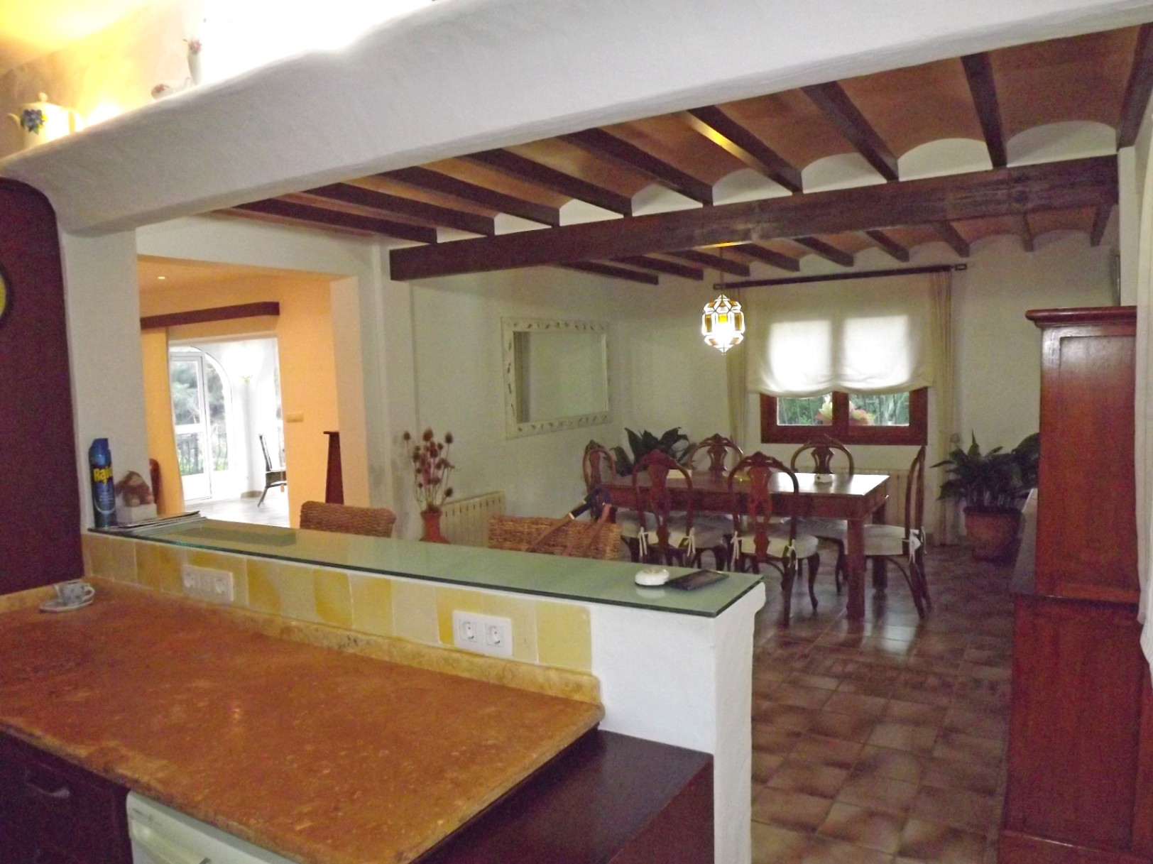 Blick von der Küche in den Essbereich mit sichtbaren dunklen Holzbalken an der Decke, Terrakotta-Elementen, Fliesenboden und einer Frühstücksbar mit Steinarbeitsplatte.