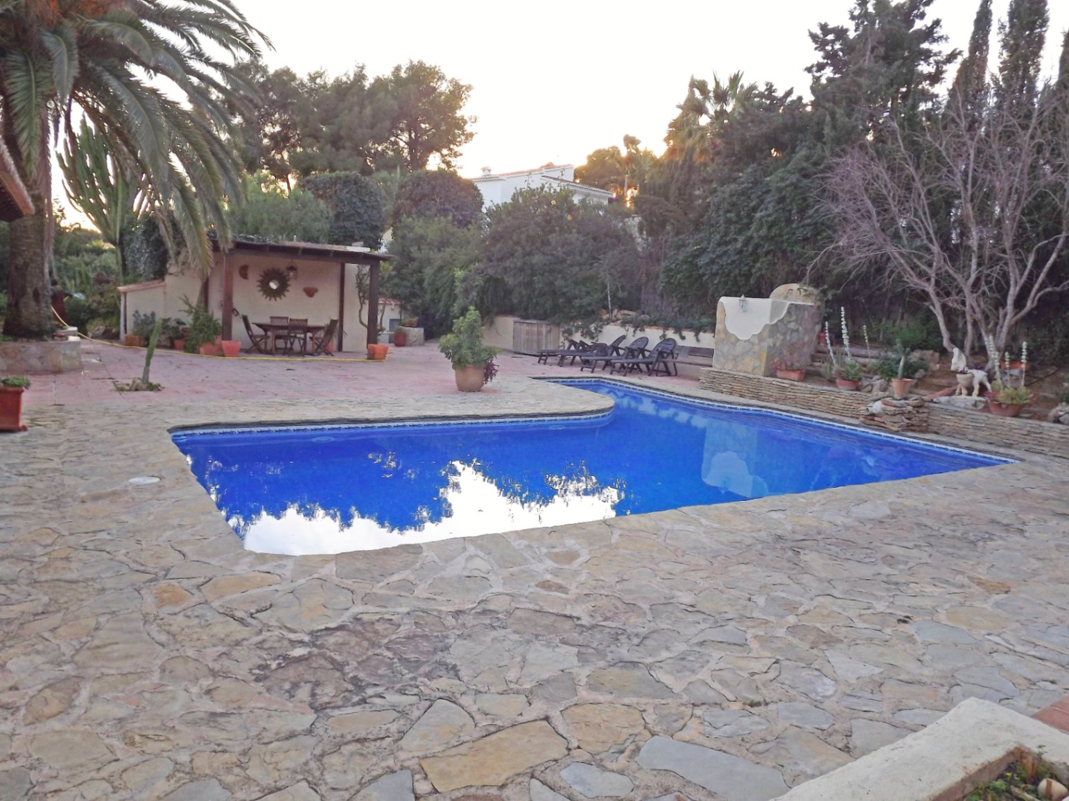 Ein 12x5m großer Pool, umgeben von einer weitläufigen Natursteinterrasse, einem überdachten Grillbereich und üppiger mediterraner Bepflanzung.