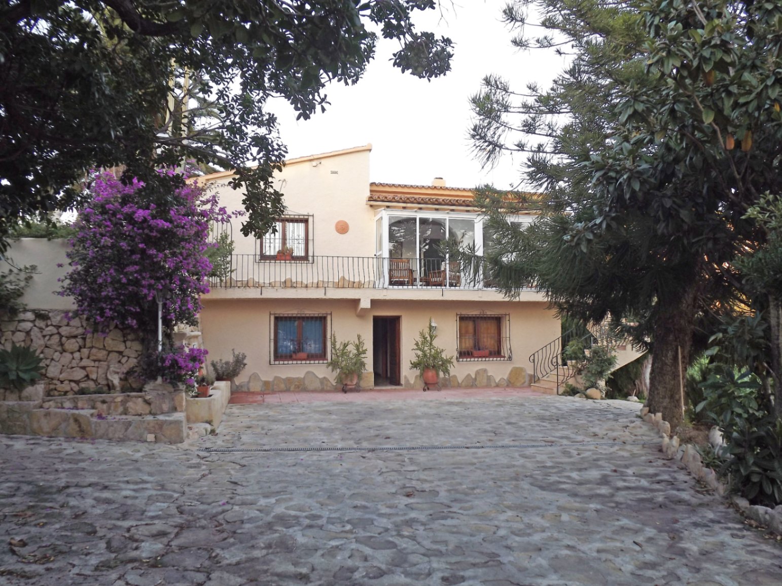 Hauptfassade der Villa mit gepflasterter Einfahrt, verglaster Oberterrasse und mediterraner Architektur, umgeben von blühenden Bougainvilleen und Pinien.
